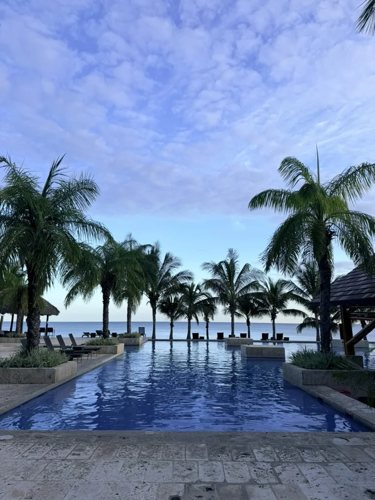 Infinity pool with blue water, surrounded by palm trees and clouds reflecting on the surface. Peaceful afternoon resort atmosphere.